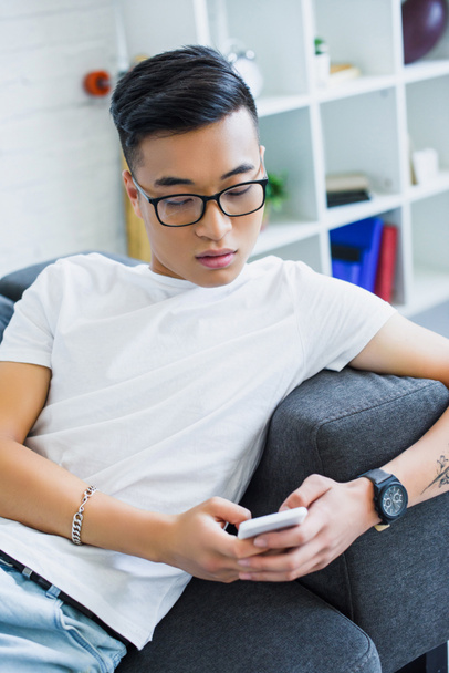 handsome asian man in glasses lying on sofa and using smartphone at home - Foto, imagen
