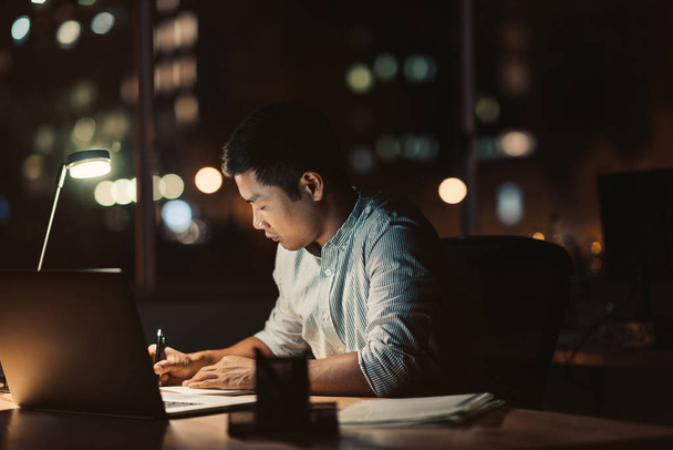 Young Asian businessman writing in a notebook while working at his desk in a dark office at night with city lights in the background - Foto, Imagem