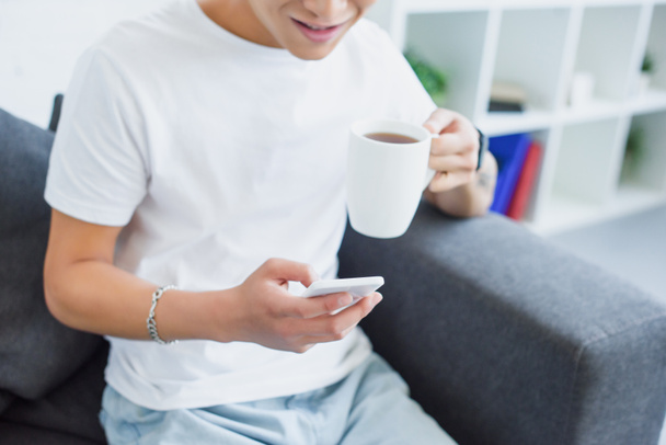 imagen recortada de hombre bebiendo té y el uso de teléfono inteligente en el sofá en casa
 - Foto, Imagen