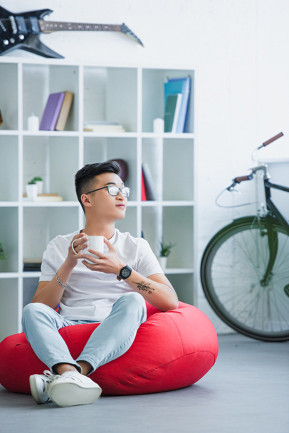 handsome asian man sitting on red bean bag chair with cup of coffee and looking away at home - Photo, Image