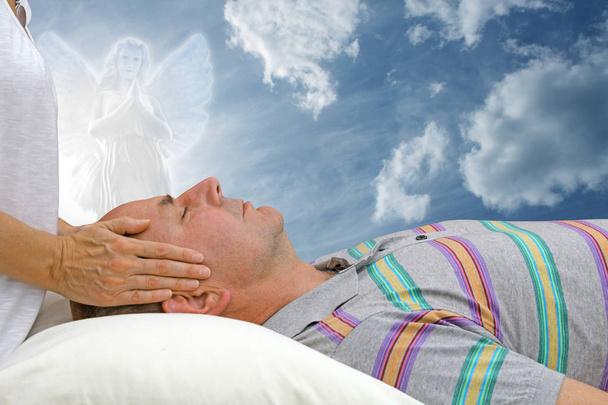Energieheilung von außen mit Schutzengel - weiblich mit Händen, die über der Stirn des Mannes schweben, liegend, gegen einen schönen Himmel mit Engel, der hinüber schaut und betet - Foto, Bild