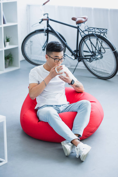 high angle view of handsome asian man sitting on bean bag chair and drinking tea at home - Photo, Image