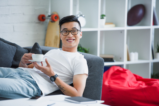 guapo feliz joven asiático hombre en gafas celebración taza de café mientras está acostado en sofá
  - Foto, Imagen