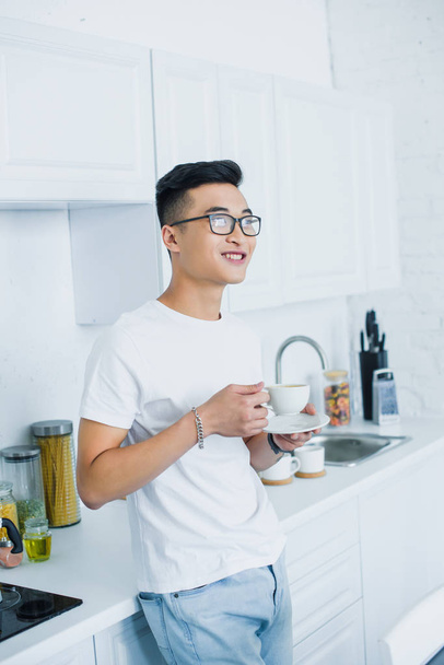 красивый улыбающийся молодой азиат в очках держит чашку кофе и смотрит на кухню
 - Фото, изображение