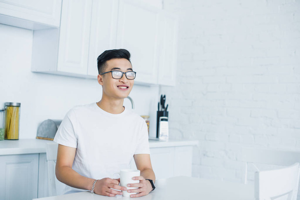 beau sourire jeune asiatique homme dans les lunettes tenant tasse de thé et regardant loin tout en étant assis dans la cuisine
  - Photo, image