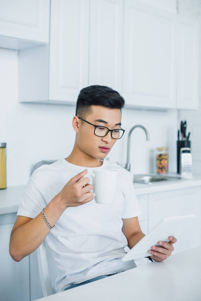 young asian man in eyeglasses holding cup and using digital tablet - Fotó, kép