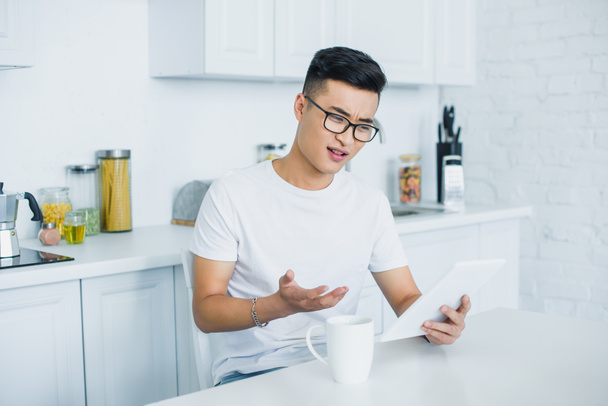 emotional young asian man in eyeglasses using digital tablet while sitting in kitchen - 写真・画像