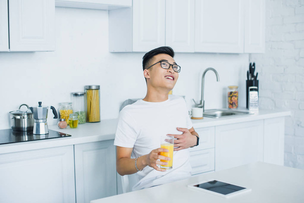 feliz joven asiático hombre celebración de vaso de jugo y mirando hacia arriba mientras sentado en cocina
 - Foto, Imagen