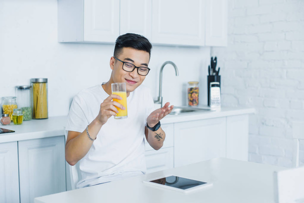 glücklicher junger asiatischer Mann mit Brille, Glas Saft in der Hand und digitalem Tablet in der Küche - Foto, Bild