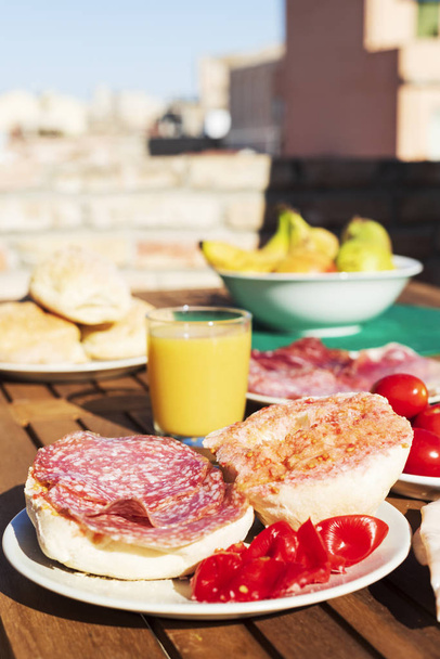 ein Sandwich mit typisch katalanischen Pa amb Tomaquet, Brot mit Tomaten, gefüllt mit Salami, auf einem Holztisch im Freien, neben einem Glas Orangensaft und einer Schüssel mit Obst - Foto, Bild