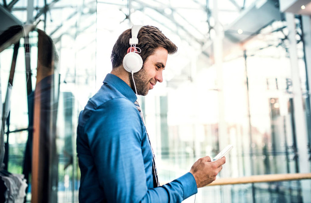 A young businessman with smartphone and headphones in a modern building. - Foto, Imagem