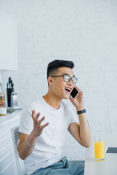 emotional young asian man talking by smartphone while sitting in kitchen - Photo, image