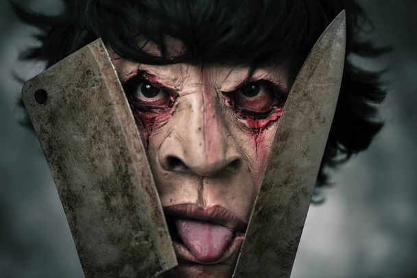 Close-up van een enge misvormde man houden een roestig en bloedige cleaver en een roestig en bloedige mes voor zijn gezicht, terwijl zijn tong uitsteekt - Foto, afbeelding