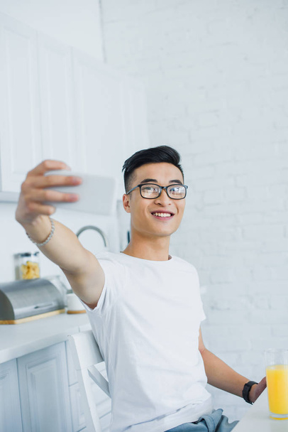キッチンでスマート フォンで selfie を取って眼鏡で微笑の若いアジア人 - 写真・画像