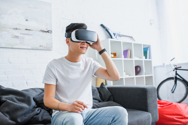 smiling young asian man using virtual reality headset while sitting on couch at home - Foto, Imagen