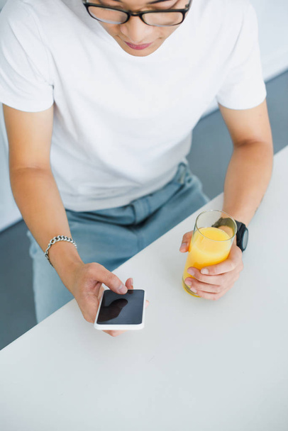 high angle view of young asian man holding glass of juice and using smartphone with blank screen - Foto, afbeelding