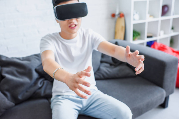 junger asiatischer Mann sitzt auf Couch und benutzt Virtual-Reality-Headset zu Hause  - Foto, Bild