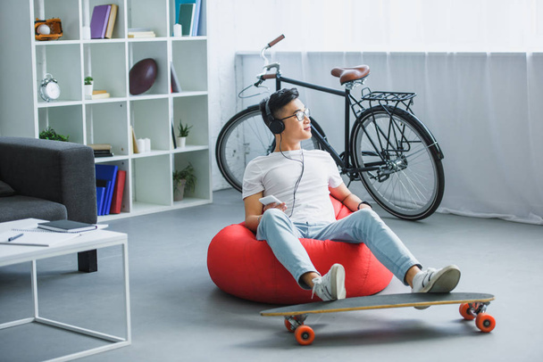 young asian man in headphones sitting in bean bag chair and using smartphone at home - Fotó, kép