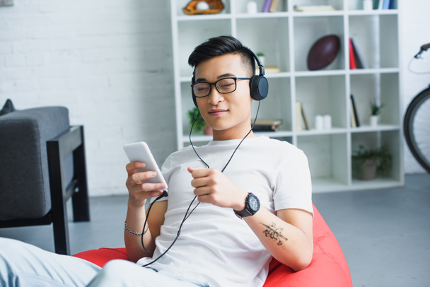 young asian man in headphones using smartphone while sitting in bean bag chair at home  - Photo, image