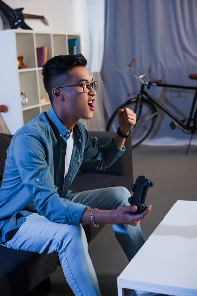 aufgeregter junger asiatischer Mann spielt Videospiel mit Joystick und gewinnt zu Hause  - Foto, Bild
