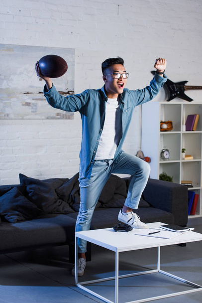 triumphing young asian man holding rugby ball and watching sport match at home - Foto, imagen