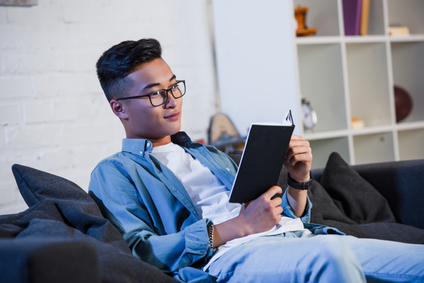 handsome smiling young man in eyeglasses sitting on couch and reading book  - Foto, Imagen