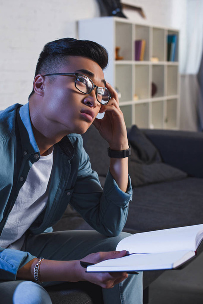 pensive young asian man in eyeglasses holding notebook and looking away at home   - Foto, Imagem
