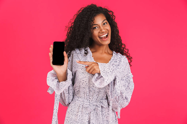 Portrait of a smiling young african woman in summer dress standing isolated over pink background, pointing at blank screen mobile phone - Fotoğraf, Görsel