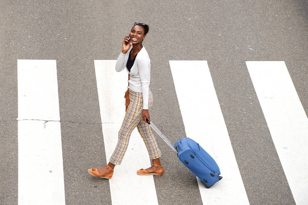 Retrato lateral de cuerpo completo de mujer afroamericana cruzando la calle con bolsas de viaje y teléfono móvil
 - Foto, Imagen