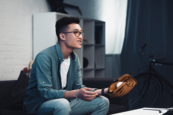 sorridente giovane uomo asiatico con guanto da baseball e palla seduta sul divano e guardando partita di sport a casa
   - Foto, immagini
