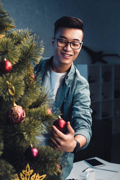handsome smiling young asian man decorating christmas tree at home  - Фото, зображення