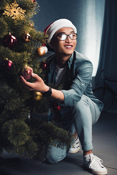 smiling asian man in santa hat decorating christmas tree and looking away  - Photo, Image