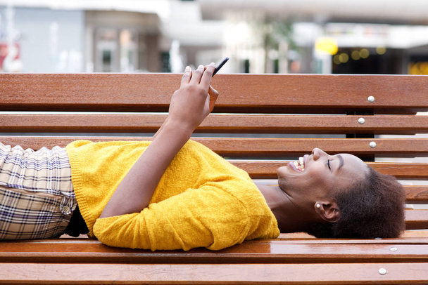 Portret van mooie jonge african american vrouw liggend op de bank kijken naar mobiele telefoon - Foto, afbeelding