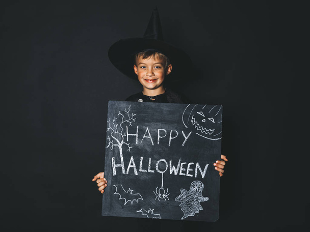 boy dressed magician costume holding happy halloween chalkboard and looking at camera in front of black background - Φωτογραφία, εικόνα