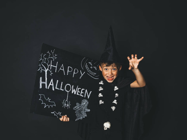 boy dressed magician costume and black cape holding happy halloween chalkboard and  showing frightening gesture while looking at camera in front of black background - Φωτογραφία, εικόνα
