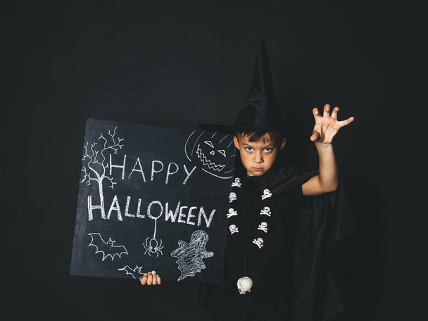 boy dressed magician costume and black cape holding happy halloween chalkboard and  showing frightening gesture while looking at camera in front of black background - Photo, Image