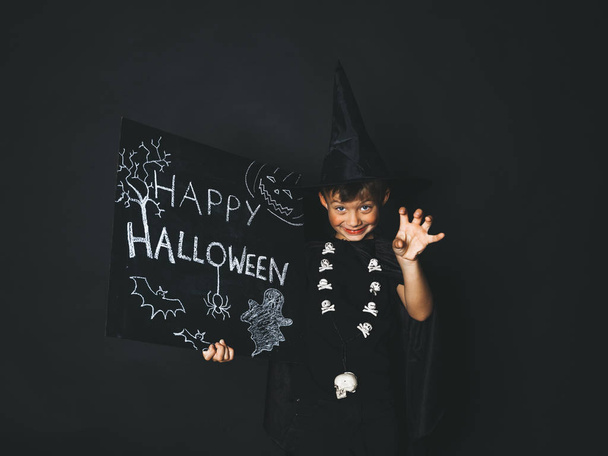 boy dressed magician costume and black cape holding happy halloween chalkboard and  showing frightening gesture while looking at camera in front of black background - Photo, Image