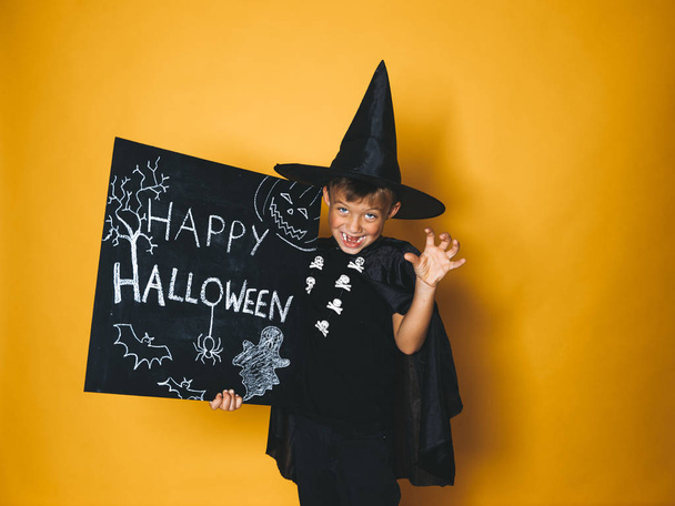 smiling little boy dressed magician hat and black cape holding happy halloween chalkboard and showing frightening gesture in front of orange background - Photo, Image