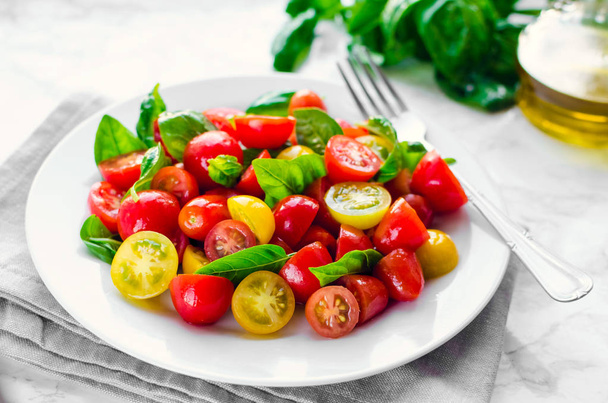 frischen hellen Tomatensalat aus roten und gelben Tomaten Kirsche mit Basilikum, Olivenöl und Balsamico Essig weißen Marmor Hintergrund. vegetarische und vegane Kost. Konzept für eine schmackhafte und gesunde Mahlzeit. - Foto, Bild