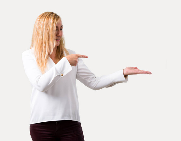 Middle age blonde woman with white shirt holding copyspace imaginary on the palm to insert an ad on isolated grey background - Photo, image