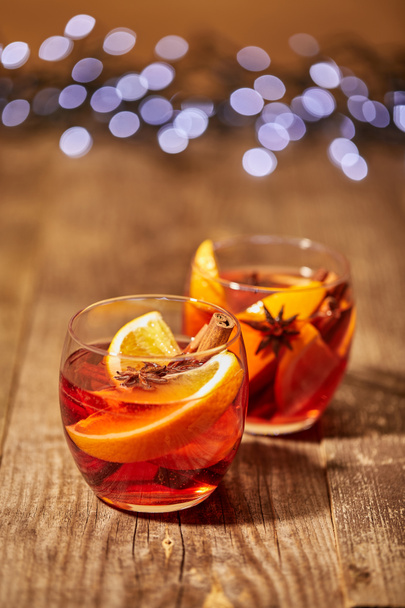 close up view of mulled wine in glasses with orange pieces and spices on wooden surface with bokeh lights on backdrop - Fotografie, Obrázek