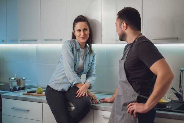 Smiling girl flirting with her boyfriend while sitting on the kitchen countertop and he is standing - 写真・画像