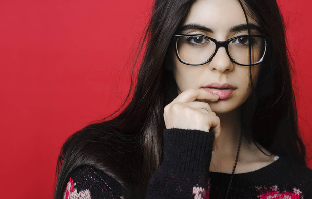 Beautiful young female in sweater and stylish glasses touching lips and looking at camera while standing on bright red background - Fotografie, Obrázek