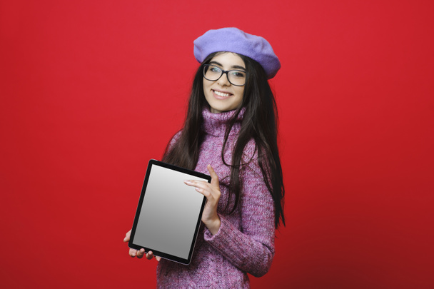 Vista lateral da jovem mulher atraente em suéter gola alta e boina elegante alegremente sorrindo e demonstrando tablet com exibição vazia enquanto está em pé no fundo vermelho brilhante
 - Foto, Imagem