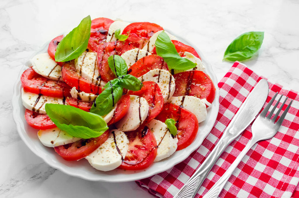 Plaat van gezonde klassieke heerlijke caprese salade met rijpe tomaten en mozzarella kaas met verse basilicum laat op witte marmeren achtergrond. Italiaanse keuken concept. - Foto, afbeelding