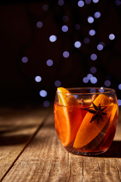 κλείνω πάνω θέα του ζεστό mulled κρασί ρόφημα με κομμάτια πορτοκαλιού και γλυκάνισο αστέρια στην ξύλινη επιφάνεια με φώτα bokeh στο παρασκήνιο - Φωτογραφία, εικόνα