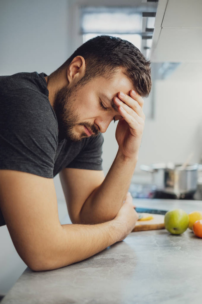 Man holding his head with his eyes closed and leaning on the kitchen countertop - Foto, Imagem