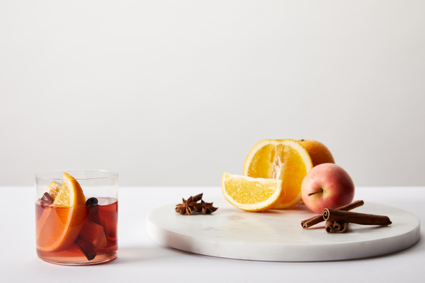 close up view of hot mulled wine in glass, spices, oranges and apple on white surface on grey backdrop - Foto, Imagen