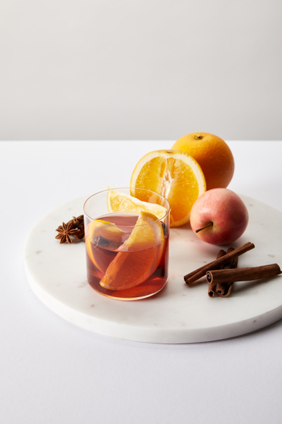 close up view of hot mulled wine in glass fresh oranges and apple on white surface on grey backdrop - Valokuva, kuva