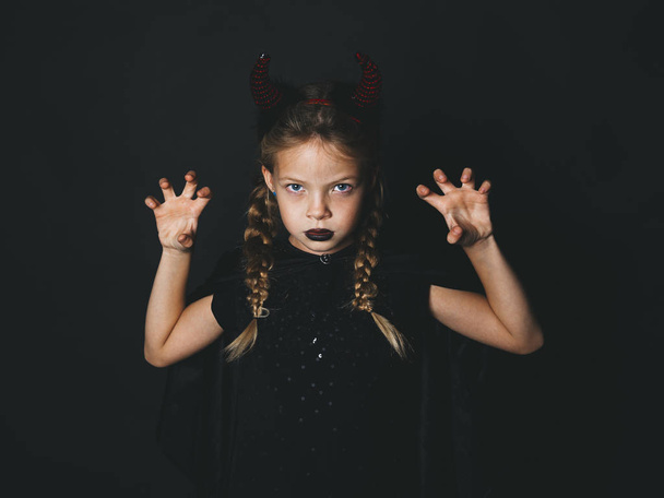 little girl in halloween devil costume with horns on head looking at camera with frightening gesture on black background  - Fotografie, Obrázek
